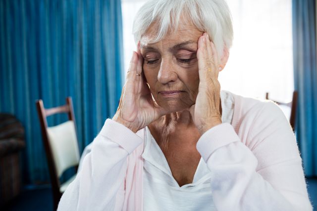 Elderly woman sitting in a retirement home, holding her head with both hands, appearing to be in pain or discomfort. This image can be used for articles or advertisements related to senior healthcare, mental health, aging, and retirement living. It is suitable for illustrating the challenges faced by the elderly, including health issues and the need for proper care and support.