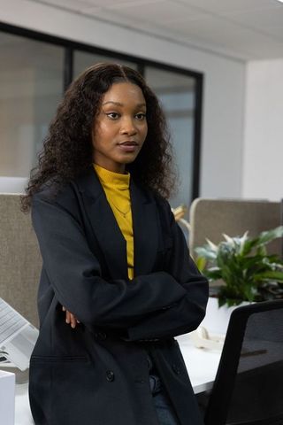 Confident businesswoman standing at modern office workstation with folded arms and blazer