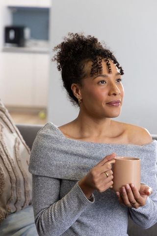 African american woman relaxing on sofa holding mug in cozy off-shoulder sweater contemplating morni