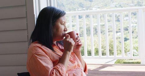 Mature asian woman relaxing on sunny balcony with ceramic mug gazing over valley
