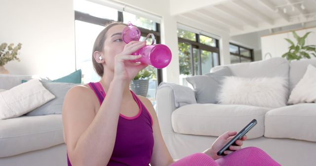 A woman stays hydrated after a workout session at home, sitting in a bright living room wearing workout attire. The image emphasizes fitness, health, hydration, and relaxation in a comfortable home environment. Perfect for marketing wellness blogs, fitness apps, gym services, health products, or articles about home-based exercise routines and healthy living tips.