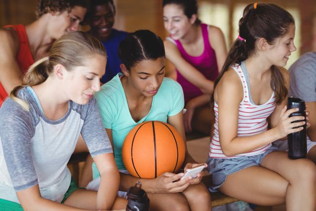 Group of high school kids sitting on a bench in a basketball court, smiling and using a mobile phone. Ideal for use in educational materials, youth sports promotions, technology in education, and social media campaigns.