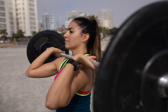 Side view close up of a strong Caucasian woman with long dark hair wearing sportswear exercising outdoors by the seaside on a sunny day, strength training holding barbells on her shoulders, concentrating.