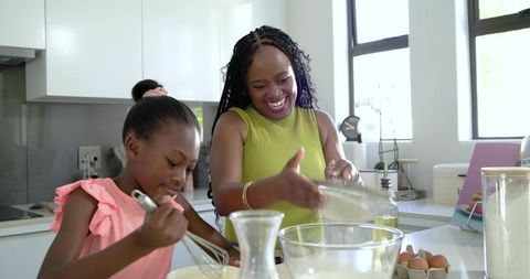 African mother and daughter baking in bright modern kitchen pouring flour and whisking