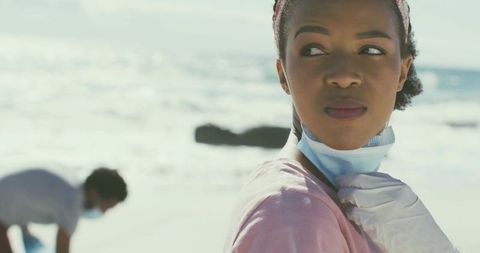 Volunteer adjusting surgical mask with gloved hand on sunny beach during coastal cleanup