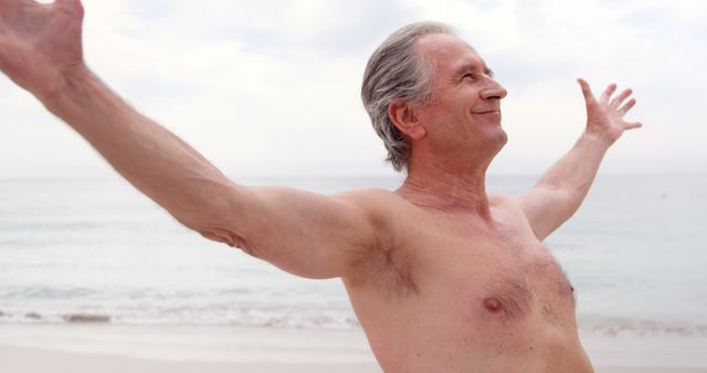 A senior man with gray hair enjoys a day at the beach, standing shirtless with his arms outstretched and a blissful expression. Perfect for themes related to aging gracefully, retirement, leisure activity, wellness, and living a joyful, carefree life.