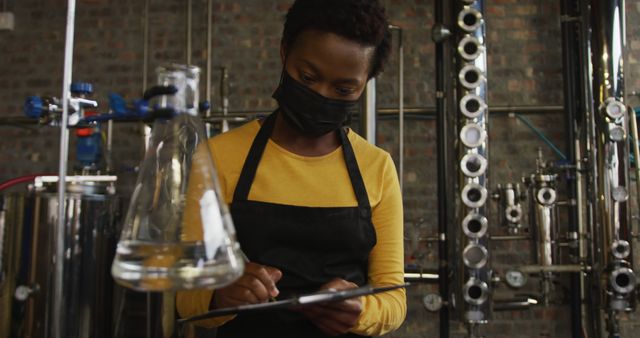 African american woman in face mask working at distillery checking equipment, writing on clipboard. work at an independent craft gin distillery business during coronavirus covid 19 pandemic.
