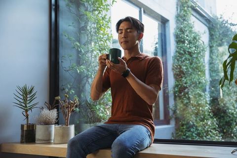Asian man sipping coffee on windowsill surrounded by indoor plants and warm natural light
