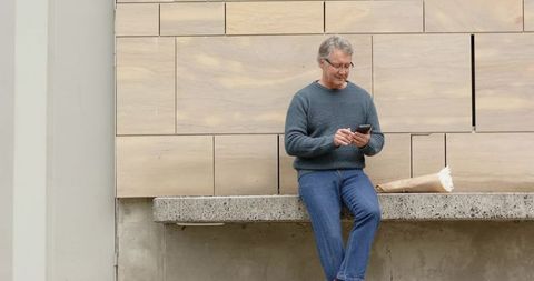 Mature man sitting on ledge checking smartphone with bouquet beside minimalist facade