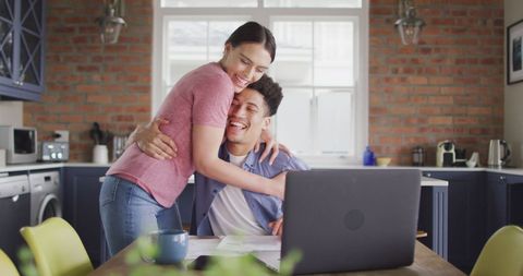 Happy Biracial Couple Embracing in Kitchen with Laptop