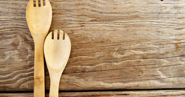 Two wooden salad forks rest on a rustic wooden table, with copy space. Their simple design and natural material suggest a homey, eco-friendly kitchen setting.