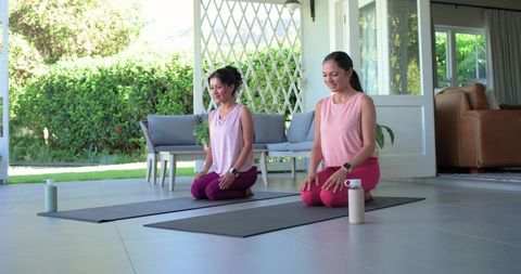 Two women practicing yoga and meditation on sunlit patio with mats and water bottles