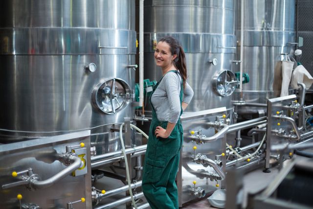 Female worker in green overalls standing confidently near large stainless steel storage tanks in a drinks production facility. Ideal for use in articles about industrial manufacturing, women in the workforce, beverage production, and factory operations.