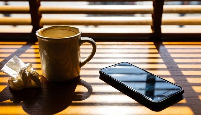 Ceramic mug catching striped sunlight on wooden table with smartphone and crumpled napkin