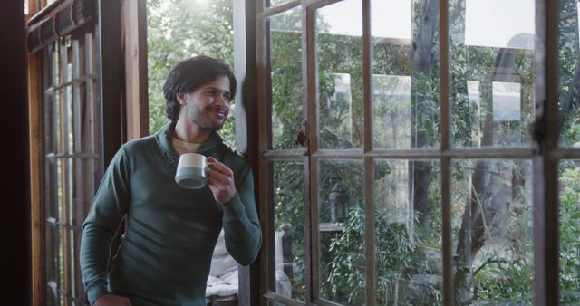 Man standing inside near large glass window holding coffee, wearing casual green sweater, enjoying view of garden greenery, appearing relaxed and content. Ideal for content focusing on relaxation, home lifestyle, solitude, morning routines, contemplation, and connection with nature.