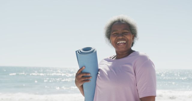 Elderly African American woman happily holding a yoga mat against a beach backdrop with a smile on her face. The image represents the joy of fitness and outdoor activities, and the concept of enjoying senior life. Perfect for promotional materials on wellness, fitness, aging, or outdoor exercises.