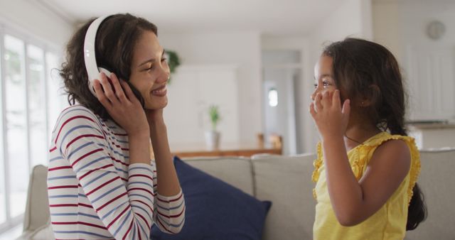 Young mother smiling while wearing headphones, sharing a joyful moment with her daughter in a bright living room. Ideal for advertisements, parenting blogs, family-oriented campaigns, and illustrating joyous family moments.