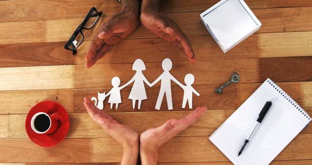 Hands encircle paper cutout figures of a family including child and pet on wooden table. Beside them are a house key, coffee cup, notepad, and pen. Useful for themes about family protection, safety, insurance, security planning, parenting, and provide a caring environment.