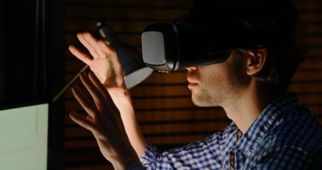 Young man experiencing virtual reality using a VR headset in a dimly lit room. This image can be used to illustrate technology, gaming, immersive experiences, and innovation. Suitable for articles, tech blogs, presentations, and promotional materials focusing on virtual reality or interactive technologies.