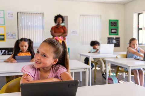 Smiling schoolgirl using tablet while African American teacher supervising classroom learning