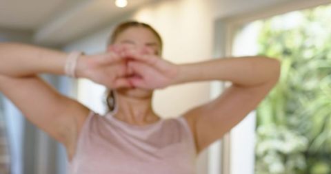 Woman covering face stretching arms wearing smartwatch by glass door with garden view