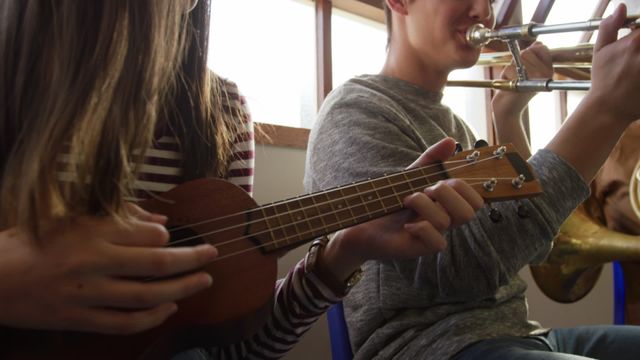 Teen male and female musicians practicing instruments during school rehearsal. Girl playing the ukulele and boy playing the trombone, both dressed casually in a well-lit room. Perfect for use in educational materials, promotions on music education, teamwork, and teen music culture representation.