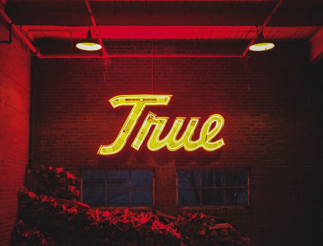 Glowing neon sign displaying the word 'True' mounted on a brick wall in an industrial warehouse setting. Ambient red lighting creates a dramatic and vintage atmosphere, making it suitable for use in projects related to nightlife, industrial design, vintage decor, or graphic design backgrounds requiring a bold and urban look.