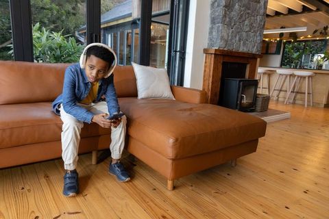 Boy wearing headphones sitting on leather sofa using smartphone in open-plan living room