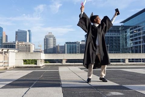 Young graduate celebrating on rooftop holding diploma and phone against city skyline