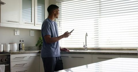 African american man using smartphone at kitchen sink with closed blinds and daylight
