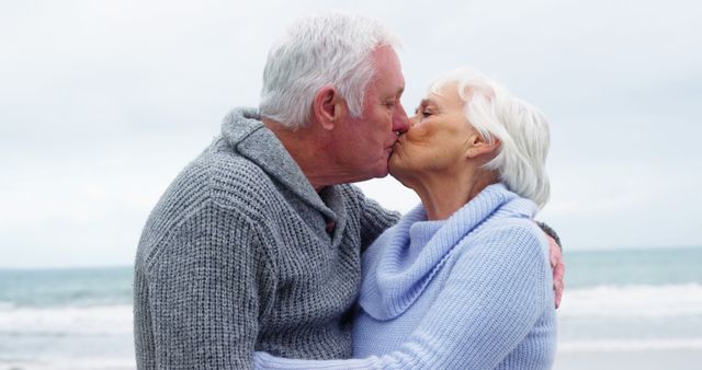 A senior Caucasian couple shares a tender kiss on a cloudy beach day, with copy space. Their affectionate moment captures a timeless love and companionship.