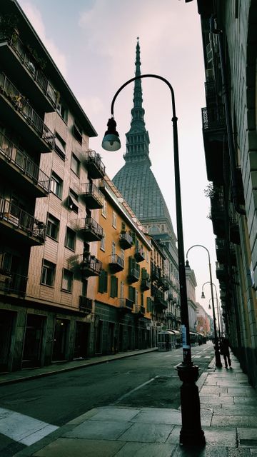 This image shows a narrow street in Turin, Italy, flanked by residential buildings and framed by the iconic silhouette of the Mole Antonelliana in the distance. Lamp posts line the street, adding a touch of urban character as dusk sets in. This image may be used in travel blogs, articles on Italian architecture, or promotional materials showcasing European cityscapes.