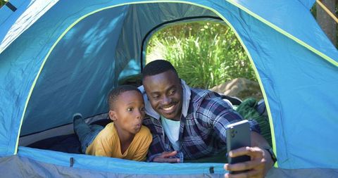 African american father and son camping together in blue tent taking selfie, sharing joyful bond