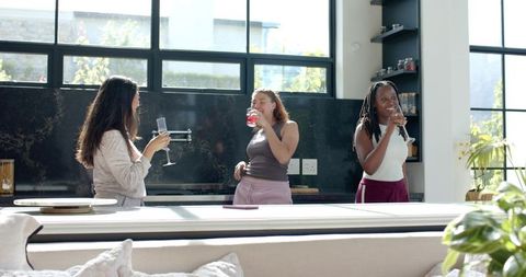 Three women laughing over drinks in sunlit modern kitchen home entertaining scene