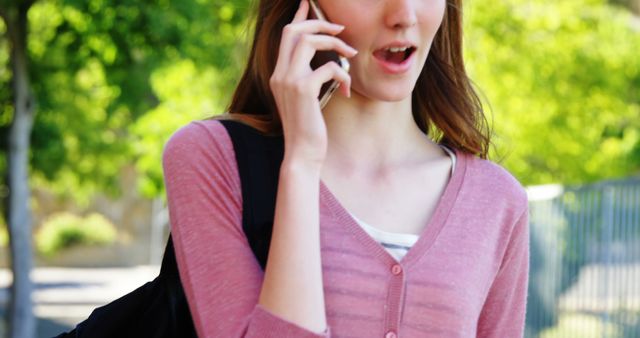 A young woman is conversing on her smartphone while walking outside. She is wearing a pink cardigan and holding a bag. This image can be used for promoting mobile communication services, lifestyle blogs, advertisements for outdoor apparel, or technology-related articles.