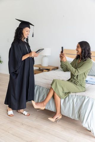 Graduate celebrating in home bedroom while sister capturing joyful moment on smartphone