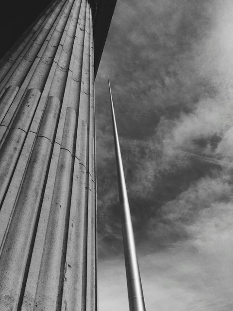 Close-up black and white perspective of an architectural column with a tall, thin tower stretching skyward. Captures the contrast and symmetry between classical and modern architectural elements, ideal for backgrounds, design inspiration, or urban-themed projects.