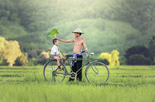 Father and son sharing a joyful moment on a bicycle in an expansive green field with scenic hills. This idyllic scene captures the essence of rural life, family bonding, and traditional values. Suitable for use in campaigns and content related to parenting, outdoor activities, countryside living, family advertisements, and cultural storytelling.