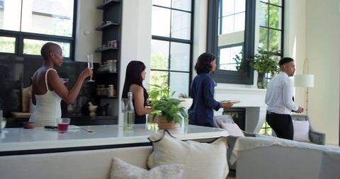Diverse friends walking through sunlit modern kitchen holding champagne flutes and plates