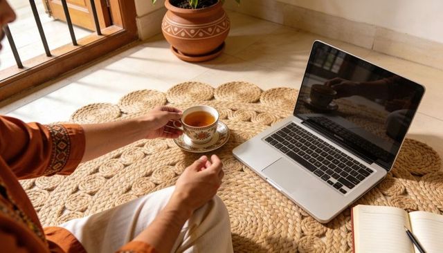 Sunlit indian man sipping tea while working on laptop on woven jute rug