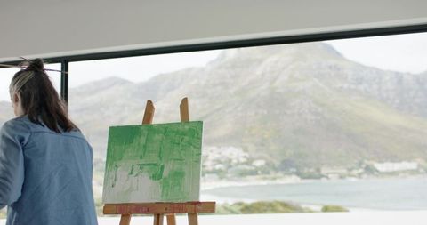 Female painter creating green abstract canvas in coastal studio with sea and mountain view