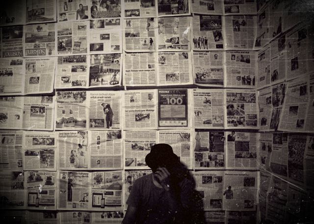 Person wearing a hat standing against a wall covered in vintage newspaper, creating a retro ambiance. This can be used for articles or presentations about history, journalism, or the evolution of printed media. It gives old-fashioned, vintage vibes perfect for themed posters, advertisements or visual stories focusing on the past.