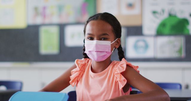 Portrait of biracial schoolgirl wearing face mask, sitting in classroom looking at camera. children in primary school during coronavirus covid 19 pandemic.