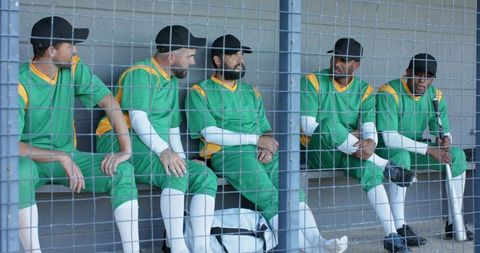 Baseball players sitting in dugout wearing green uniforms behind chain-link fence