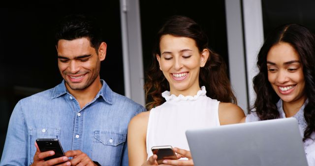 Young professionals are standing together in an office environment, engaged with their smartphones and laptop. All are smiling, indicating enjoyment and camaraderie in a modern workplace. This image can be used to depict teamwork, digital communication, technology usage, and a positive work environment. Ideal for articles, advertisements, or website content focusing on business, technology, and young professional culture.