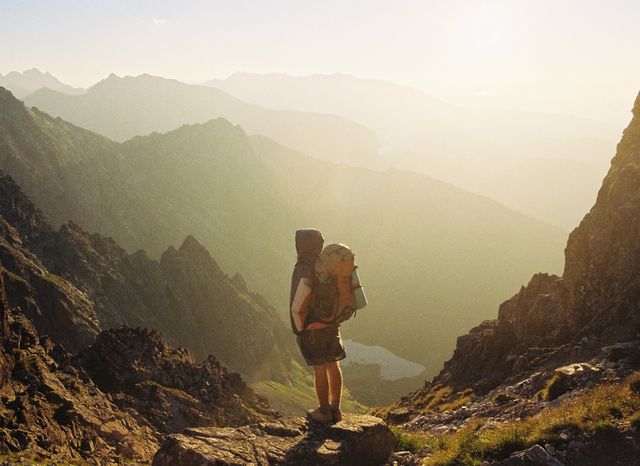 Backpacker stands on rocky terrain while admiring the breathtaking sunrise over mountain range. Perfect for use in travel and adventure marketing materials, nature documentaries, outdoor gear advertisements, and articles about solo travel and hiking.