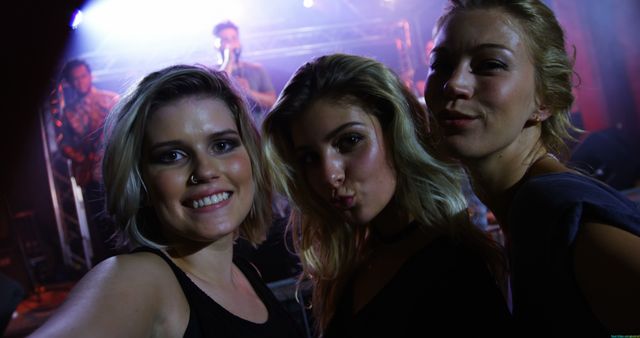 Three young Caucasian women are taking a selfie at a concert, with vibrant stage lights illuminating the background. Their joyful expressions capture the lively atmosphere of the music event they are attending.
