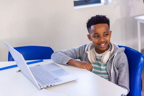 African american boy studying online on laptop with headphones in bright modern classroom