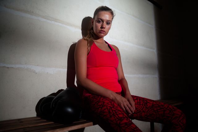 This image shows a sad woman sitting on a bench in a fitness studio, wearing a red sports outfit. She appears to be resting after a workout, with boxing gloves placed beside her. This image can be used for articles or advertisements related to fitness, mental health, emotional well-being, or sports training.