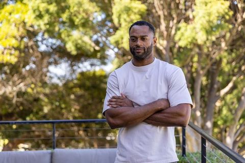 Contemplative mature african man standing on balcony with arms crossed, wearing light tee